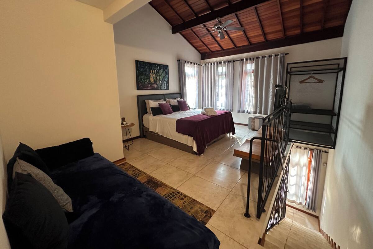 A bedroom with a bed, a nightstand, a ceiling fan, a window with curtains, and a staircase.