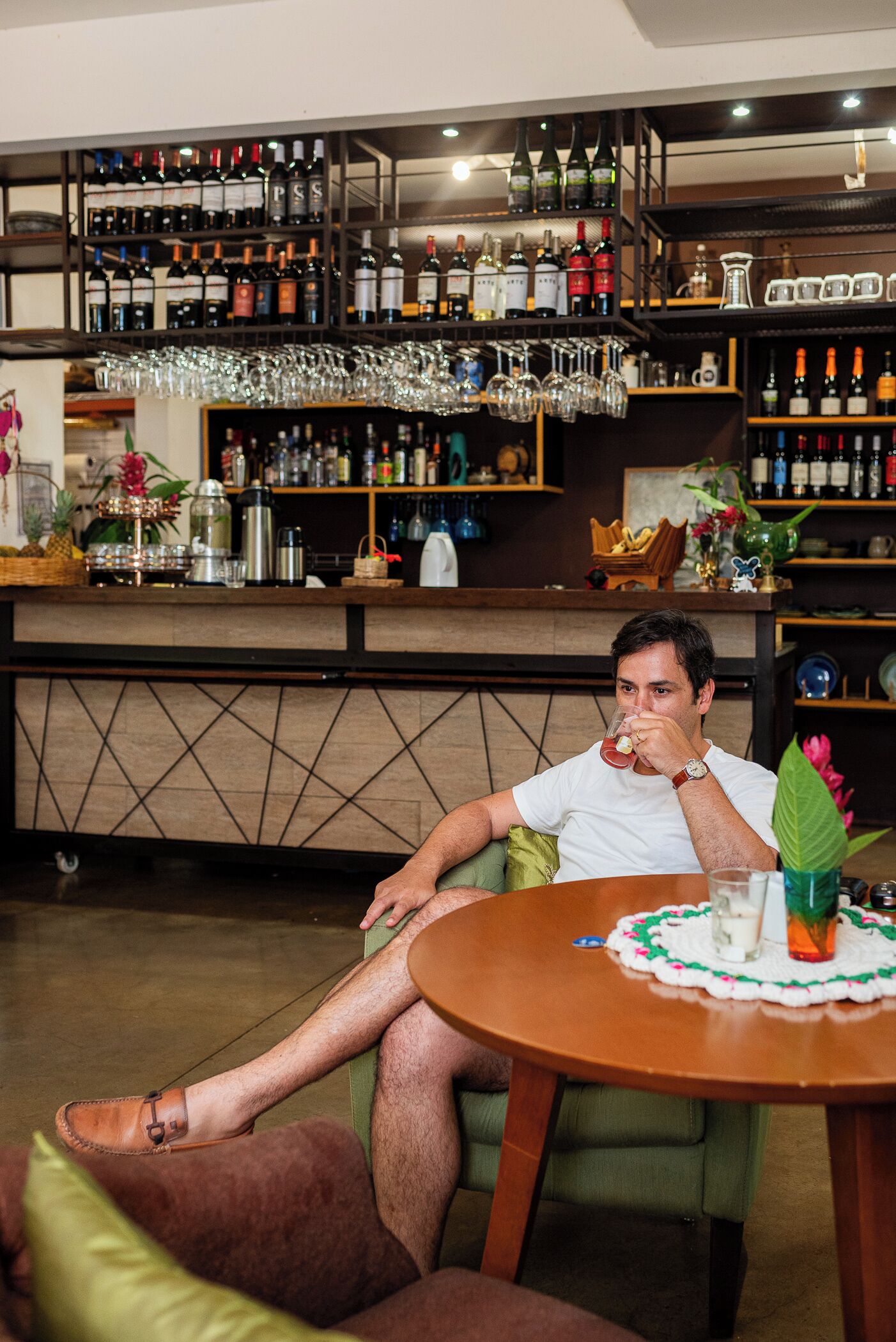 Um homem sentado a uma mesa redonda com uma bebida, num espaço com um bar repleto de garrafas e copos diversos.