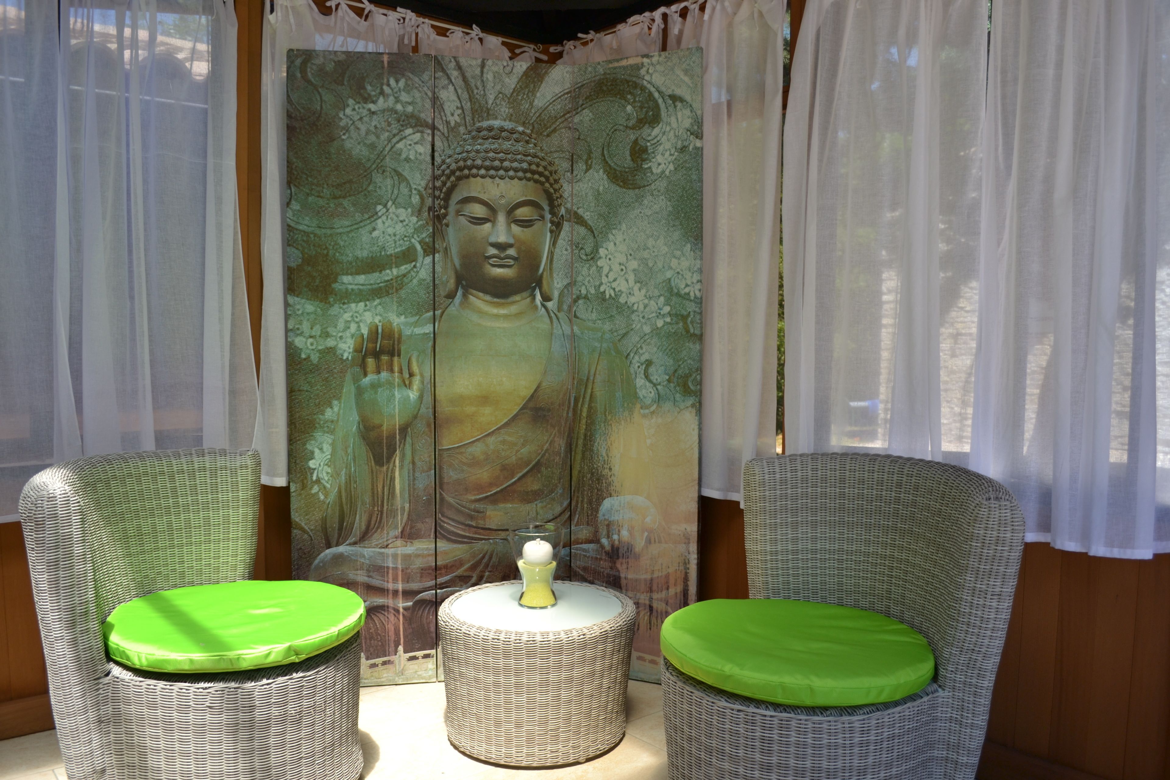 A room with two wicker chairs with green cushions, a small round table with a green cushion, and a Buddha painting on the wall.