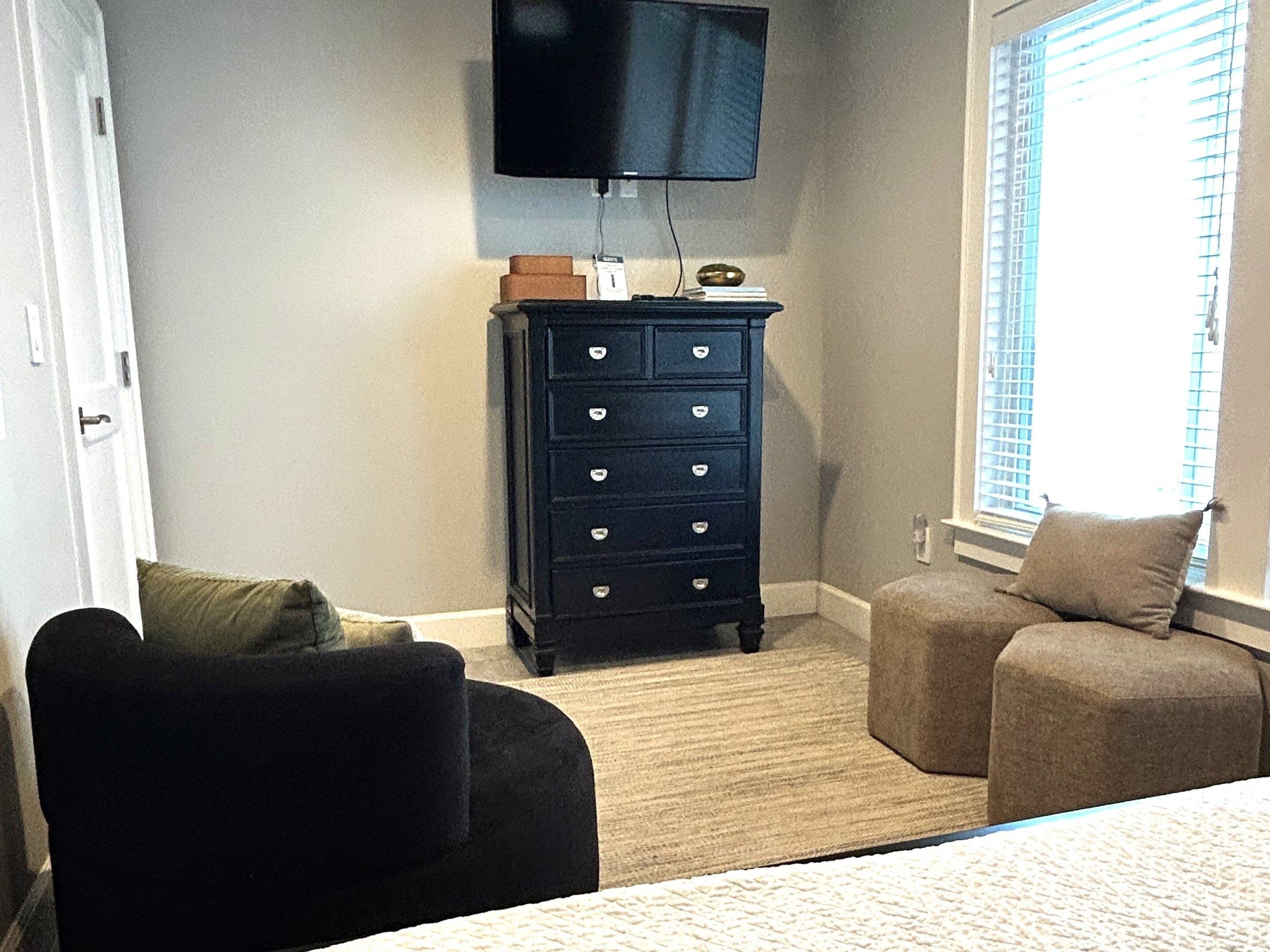A bedroom with a bed, a black chair, a small ottoman, a dresser, and a television on the wall.