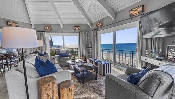 A spacious living room with a large window overlooking the beach, furnished with a grey sofa, armchairs, and a coffee table.