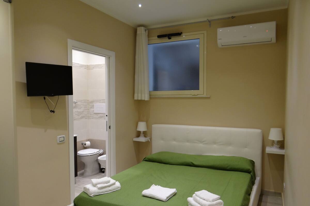 Una habitación pequeña y limpia, con colcha verde, cabecero blanco y dos lámparas de noche. Un televisor de pantalla plana está montado en la pared y hay una ventana con cortina.