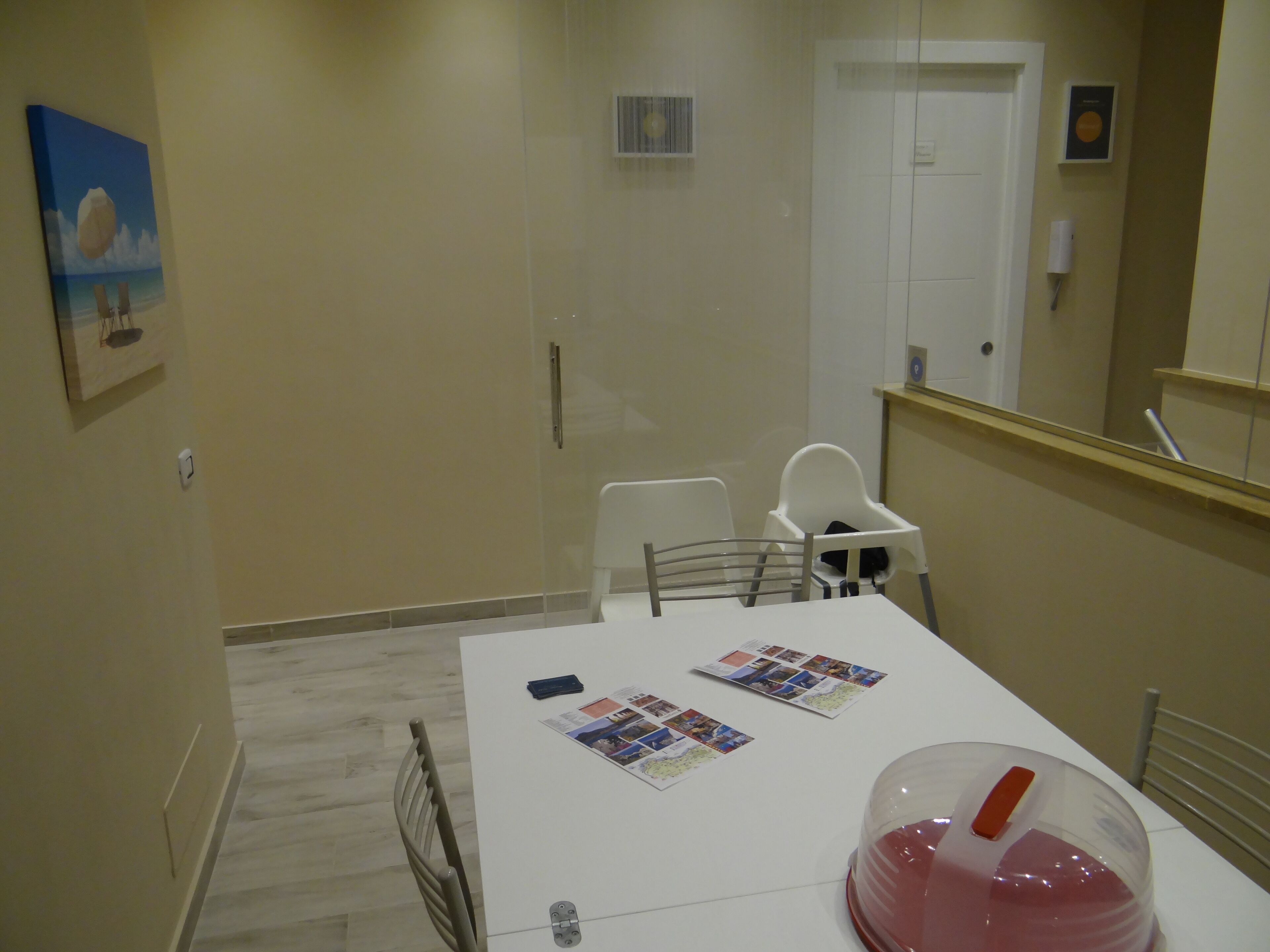 Un pequeño comedor con una mesa blanca, dos sillas y una trona para bebé. Hay fotos sobre la mesa y un contenedor de plástico grande a la derecha.