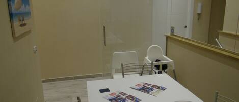 Un pequeño comedor con una mesa blanca, dos sillas y una trona para bebé. Hay fotos sobre la mesa y un contenedor de plástico grande a la derecha.