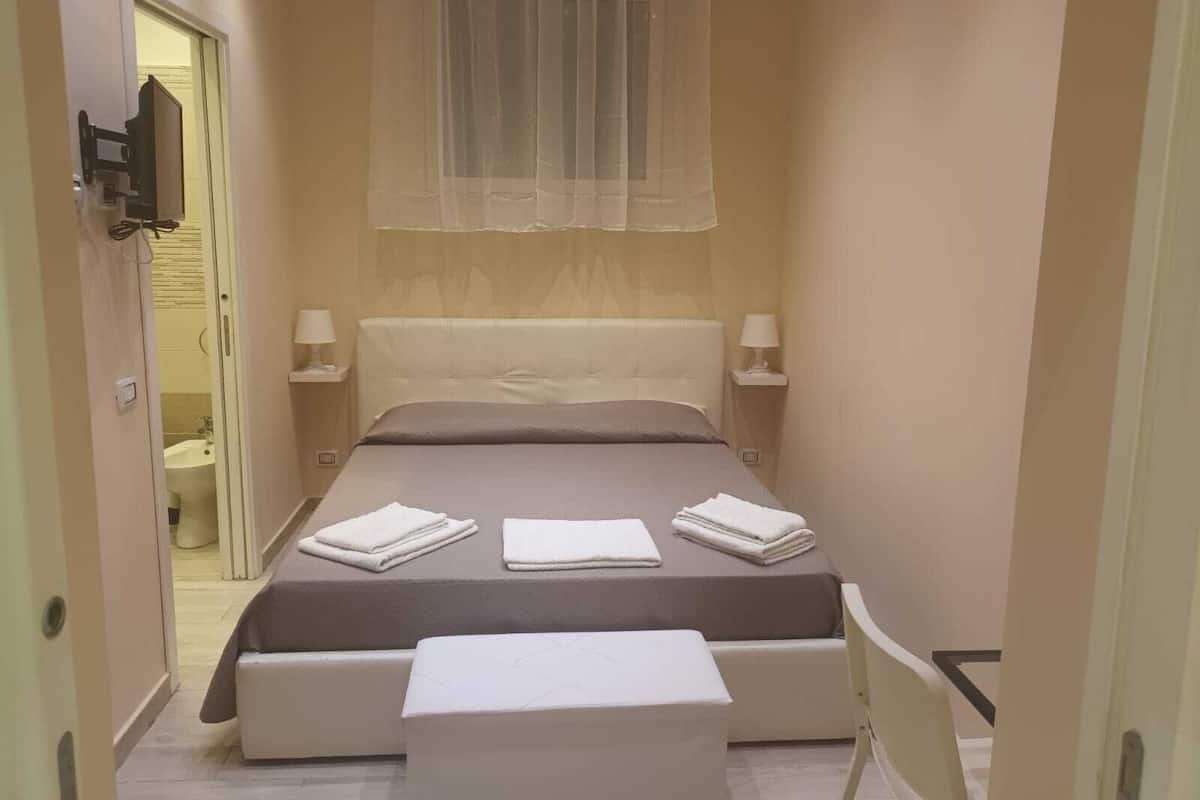 Habitación de hotel estrecha con cama, lámparas de noche, ventana con cortinas translúcidas, una mesita y una silla.