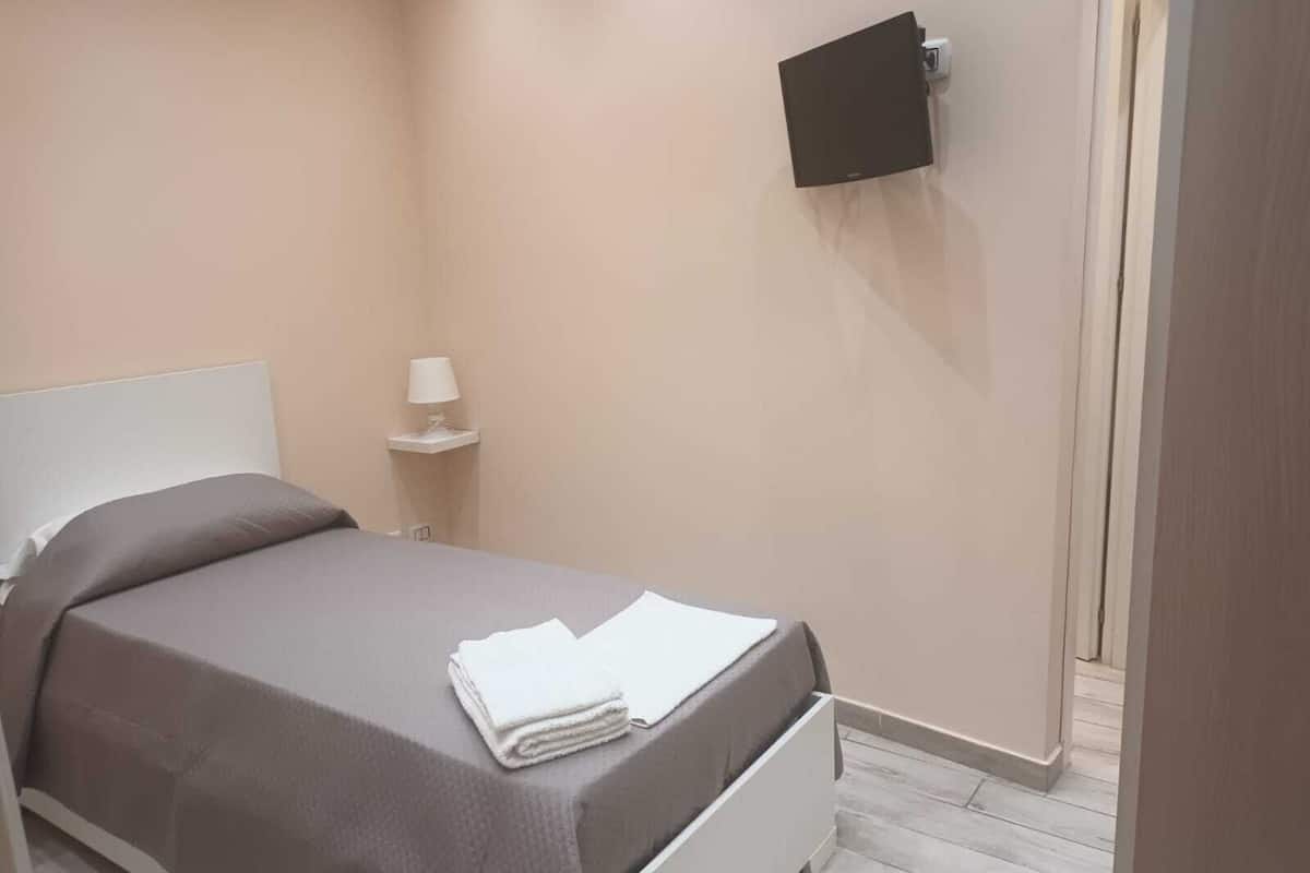 Una cama bien hecha con cabecera blanca, una mesita de noche con lámpara y un televisor de pantalla plana montado en la pared.