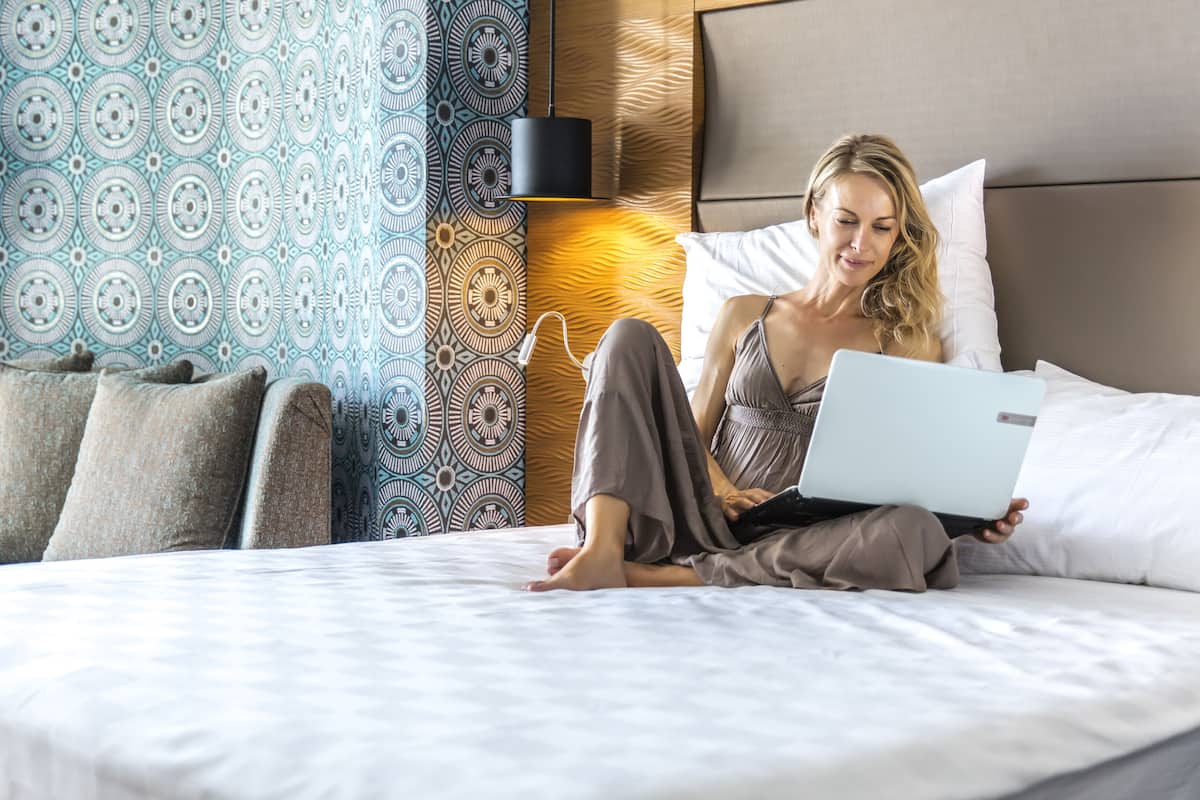 Eine Person sitzt auf einem Bett mit einem Laptop, einer Wand mit Muster und einem Kopfteil.