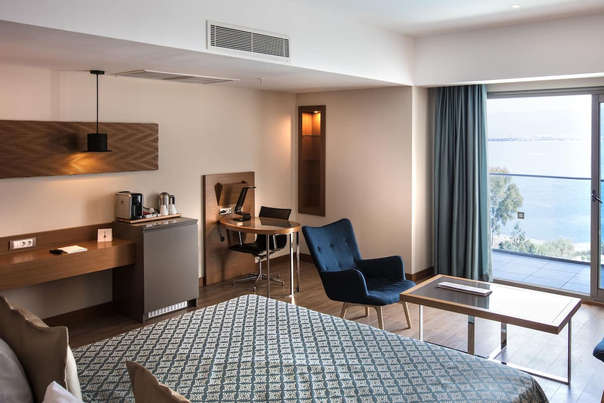 Ein Hotelzimmer mit einem Bett, einem Schreibtisch, einem Sessel, einem kleinen Tisch und einem Fernseher. Ein Fenster bietet Blick auf das Meer.