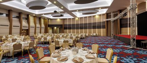 Un gran salón de banquetes con mesas redondas dispuestas para comer, un escenario con fondo rojo y una alfombra estampada.
