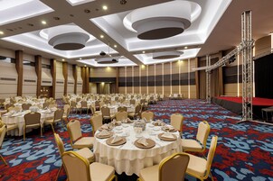 Una grande sala da banchetto con tavoli rotondi apparecchiati per la cena, un palco con uno sfondo rosso e una tappezzeria decorata.