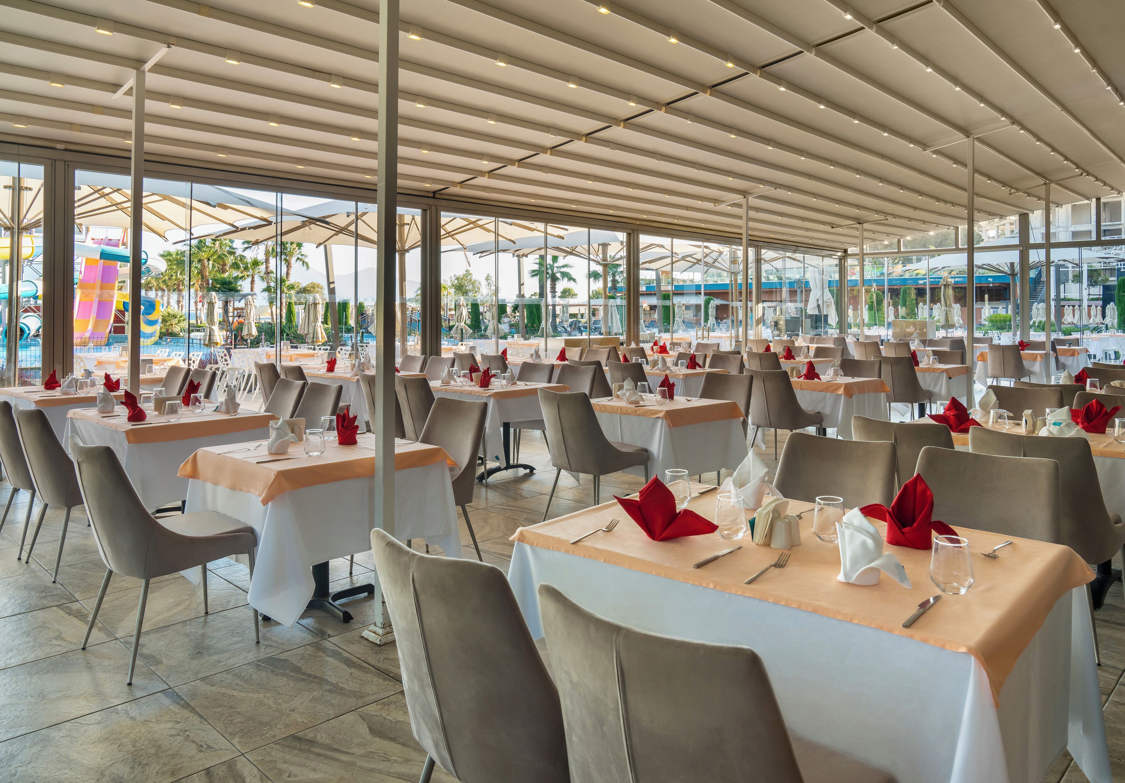 A spacious outdoor dining area with multiple tables and chairs, large windows, and a view of a water park.