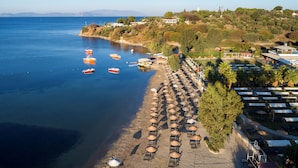 A beach with rows of sunbeds and umbrellas, a clear blue sea, and boats anchored nearby. - Liberty Kuşadası - All inclusive (Kusadasi)