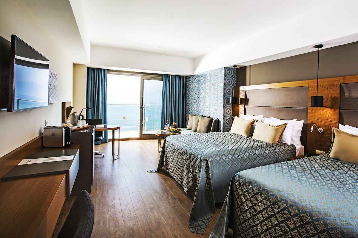 Ein Hotelzimmer mit zwei Betten, einem Schreibtisch, einem Sessel, einem Fernseher und Blick auf das Meer.