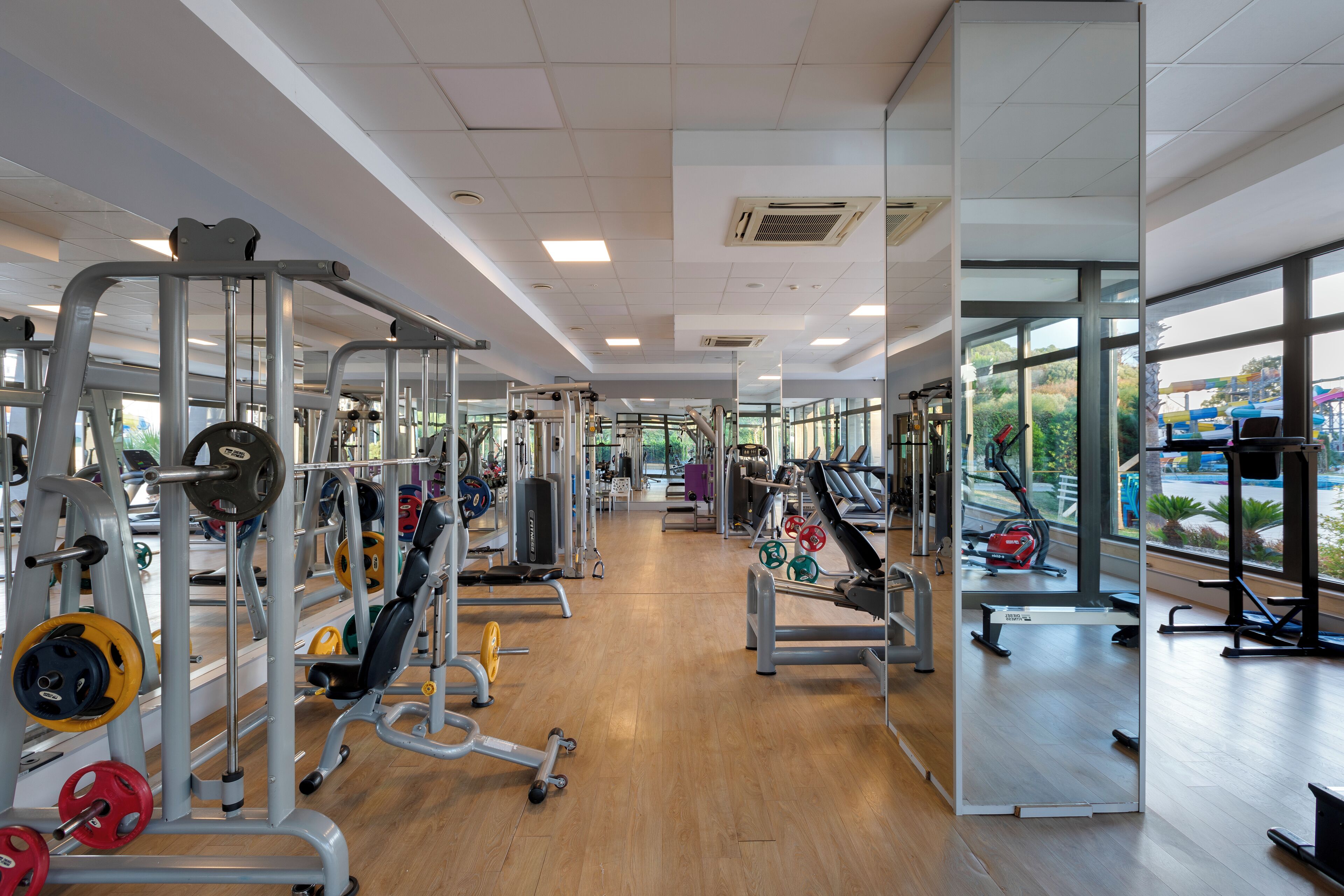 Een sportschool met diverse fitnessapparaten, grote ramen en een houten vloer.