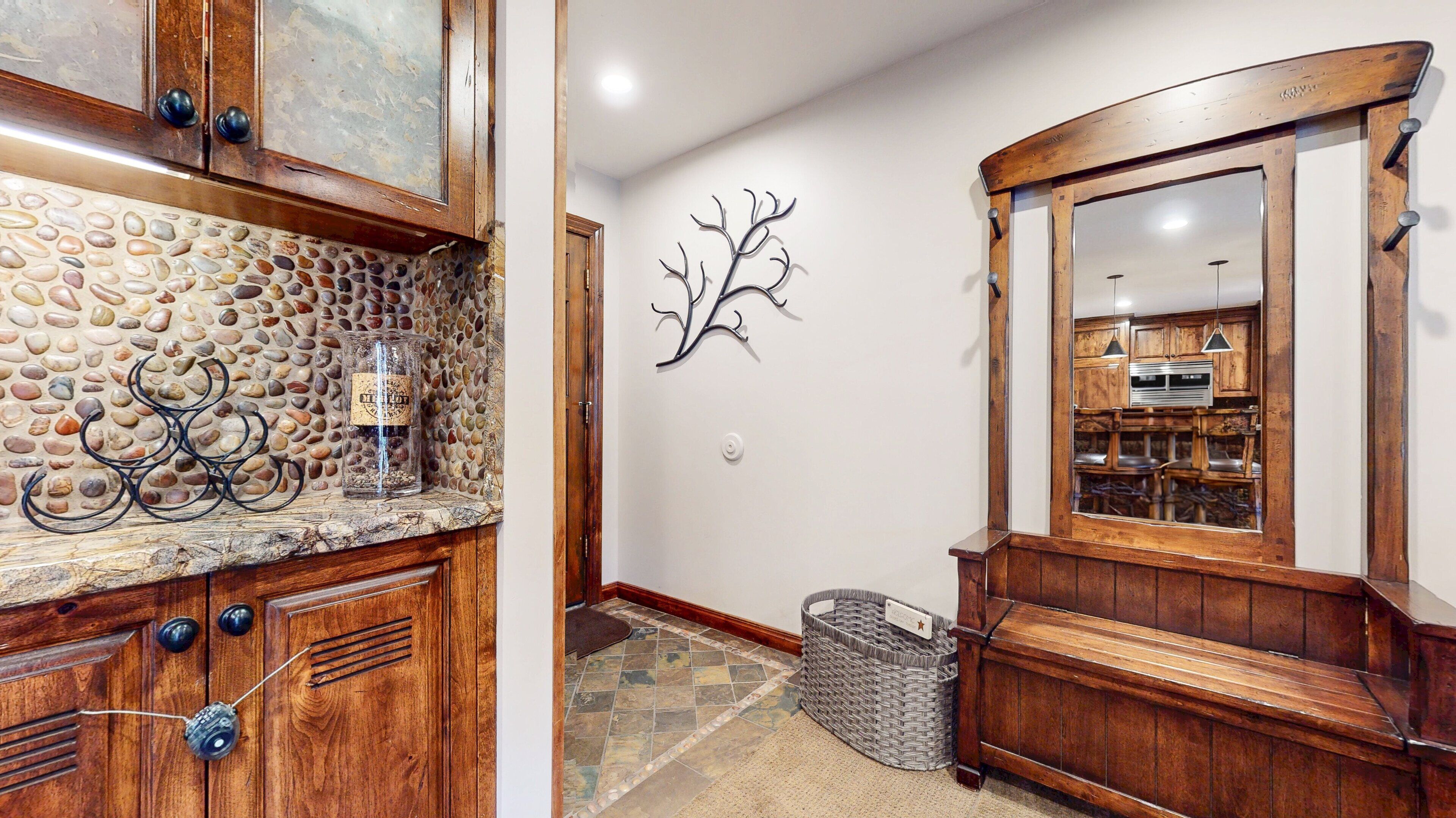 A wooden hallway with a stone mosaic backsplash, a metal tree sculpture, and a large mirror reflecting a kitchen area.