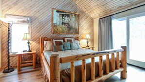 Um quarto com teto de madeira, uma cama grande com estrutura de madeira, duas mesas de cabeceira com luminárias, uma janela ampla com cortinas e um quadro acima da cama.
