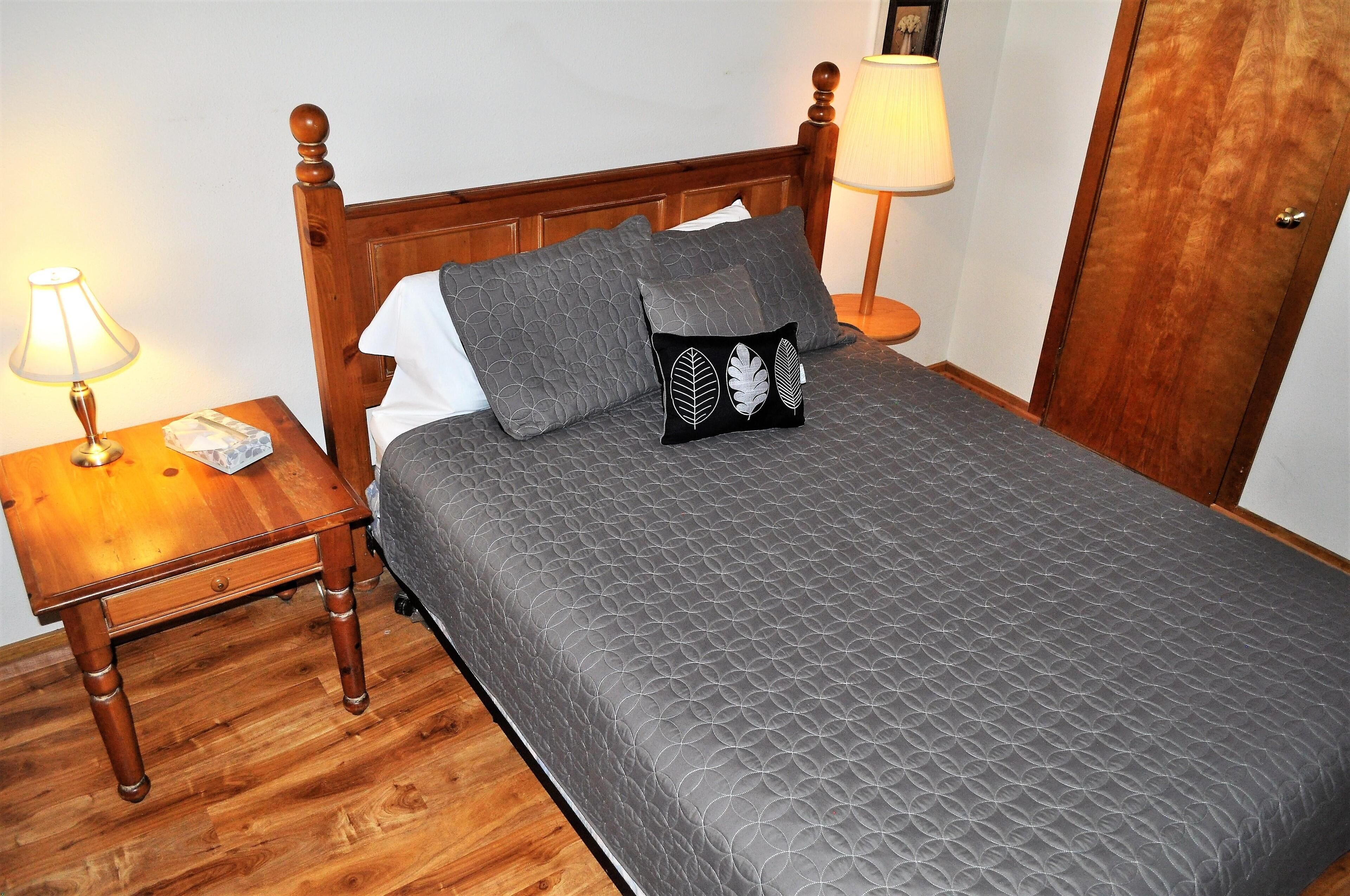 Un lit bien fait, avec une literie grise et deux oreillers, une table de chevet en bois avec une lampe, et une porte en bois à l’arrière-plan.