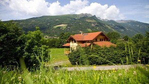 Une maison avec un toit rouge, entourée d’une végétation luxuriante et avec des montagnes en arrière-plan.