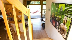 Un escalier en bois mène à une pièce où se trouvent un grand tableau représentant Bouddha, un hamac et une étagère en bois remplie de livres et d’objets décoratifs.