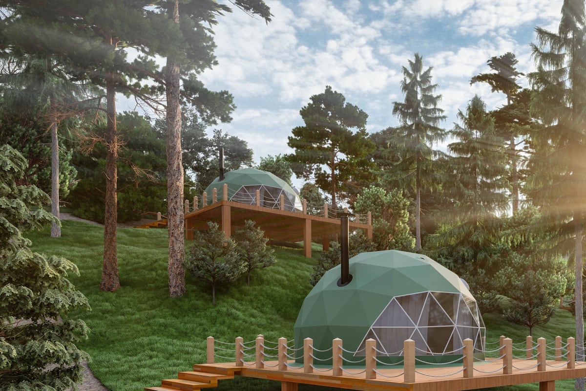 A geodesic dome structure surrounded by a wooden deck and a rope barrier, set in a forested area with a clear sky.