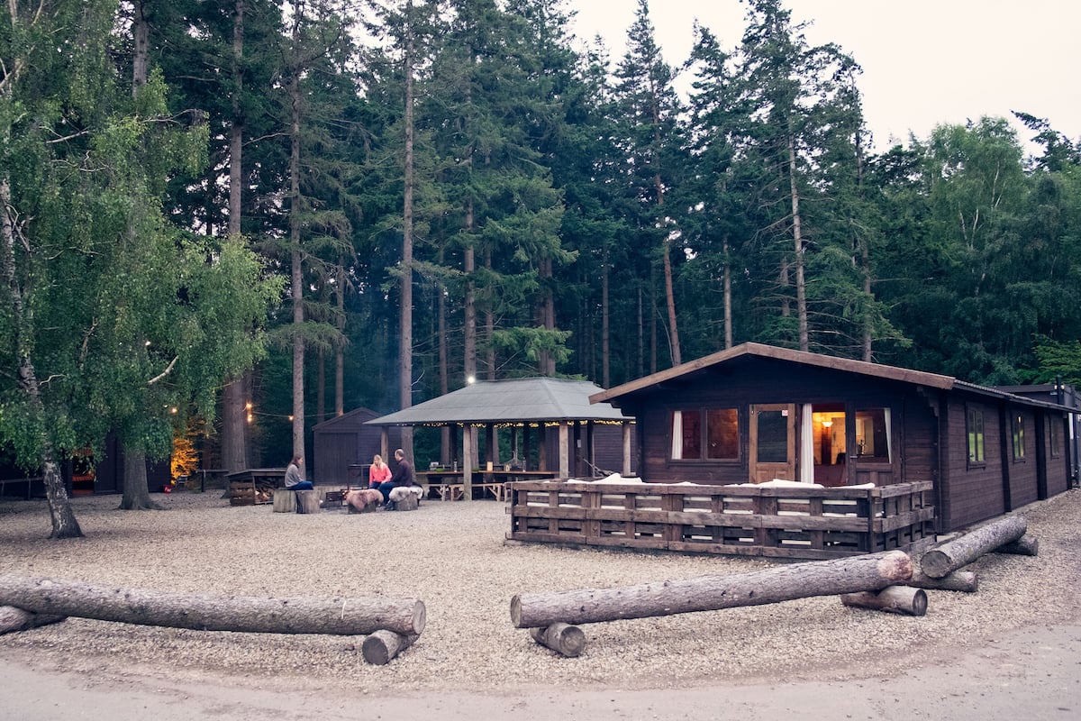 A rustic cabin with a wooden deck and a covered picnic area, surrounded by dense forest.
