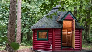 Eine kleine, rote Holzhütte mit grünem Fenster und Tür, umgeben von einem Wald.