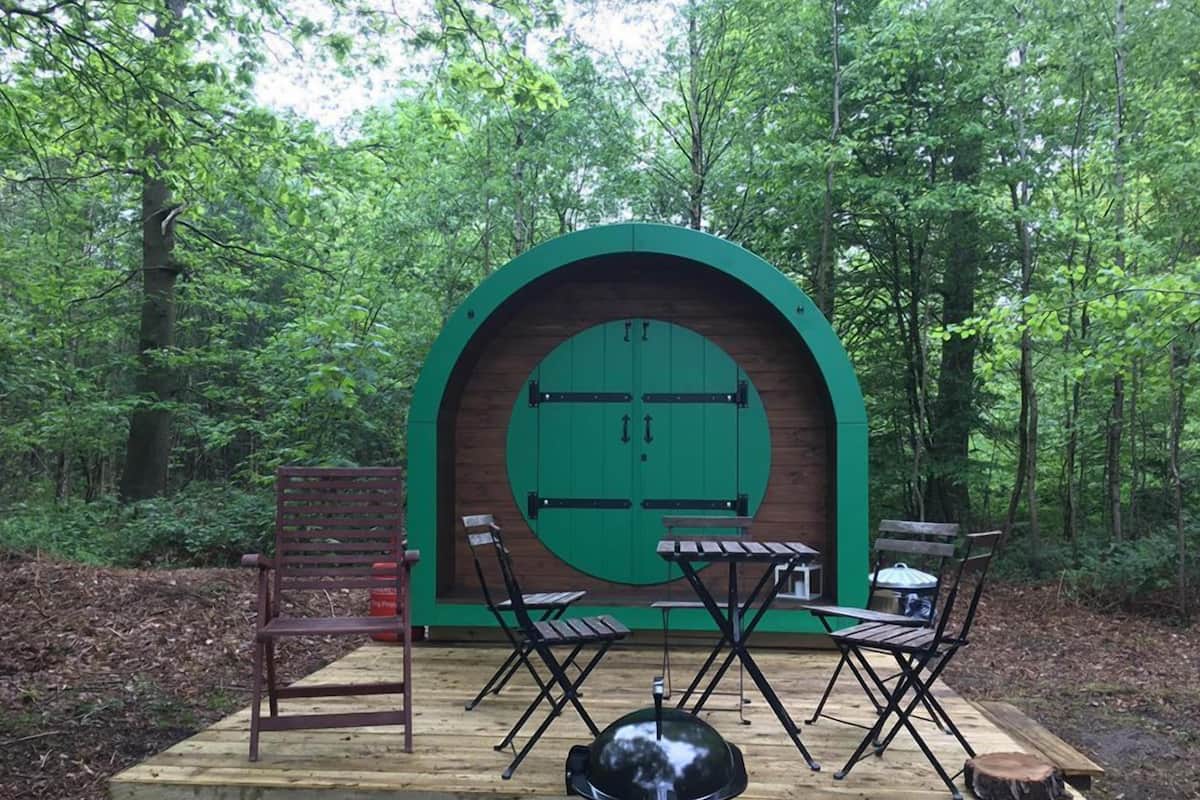 A rustic wooden deck with a green archway, two folding tables, and chairs, surrounded by a forest.