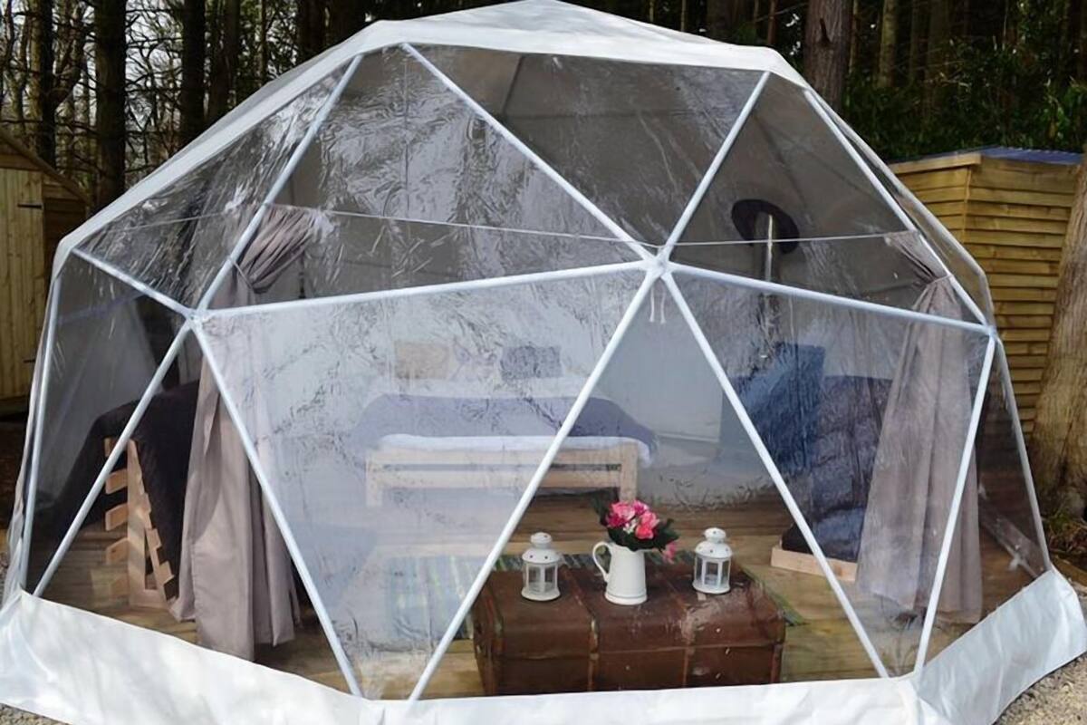 A geodesic dome structure with a transparent covering, a wooden bench, and a table with lanterns and a vase.