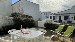 Un patio con una mesa dispuesta para dos personas, rodeado de sillas de plástico blanco, una botella de vino y copas.