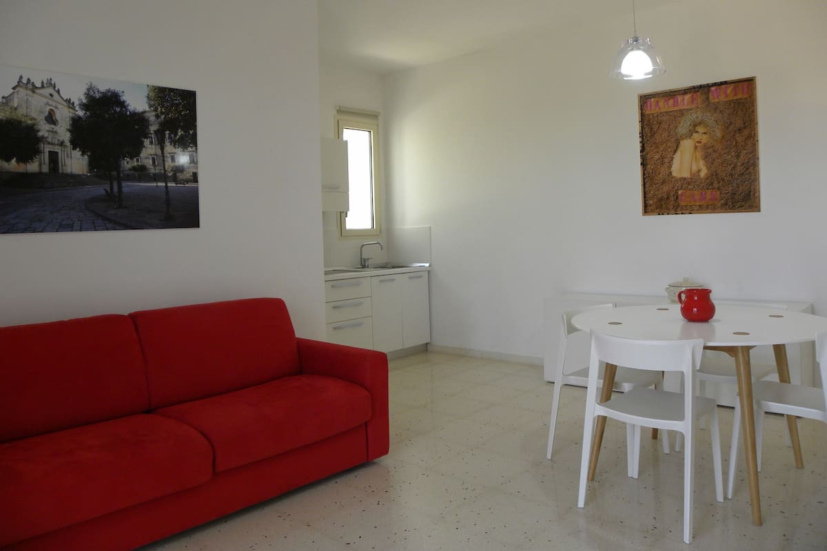 Ein Zimmer mit einem roten Sofa, einem weißen Esstisch mit Stühlen, einer Küchenzeile und einem Bild an der Wand.