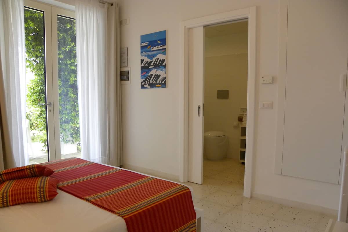 Ein Schlafzimmer mit Bett, ein durchgehender Blick ins Badezimmer und ein Fenster mit Vorhängen.