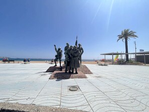 Uma praça pública com uma estátua de soldados, bancos e vista ampla para o mar.