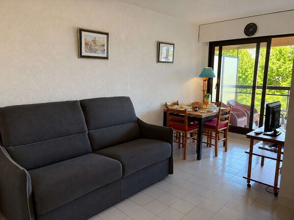 A living room with a sofa, dining table, chairs, a TV, and a window with a view. - Seaside studio cabin, 4 pers, terrace, quiet residence (Fréjus)