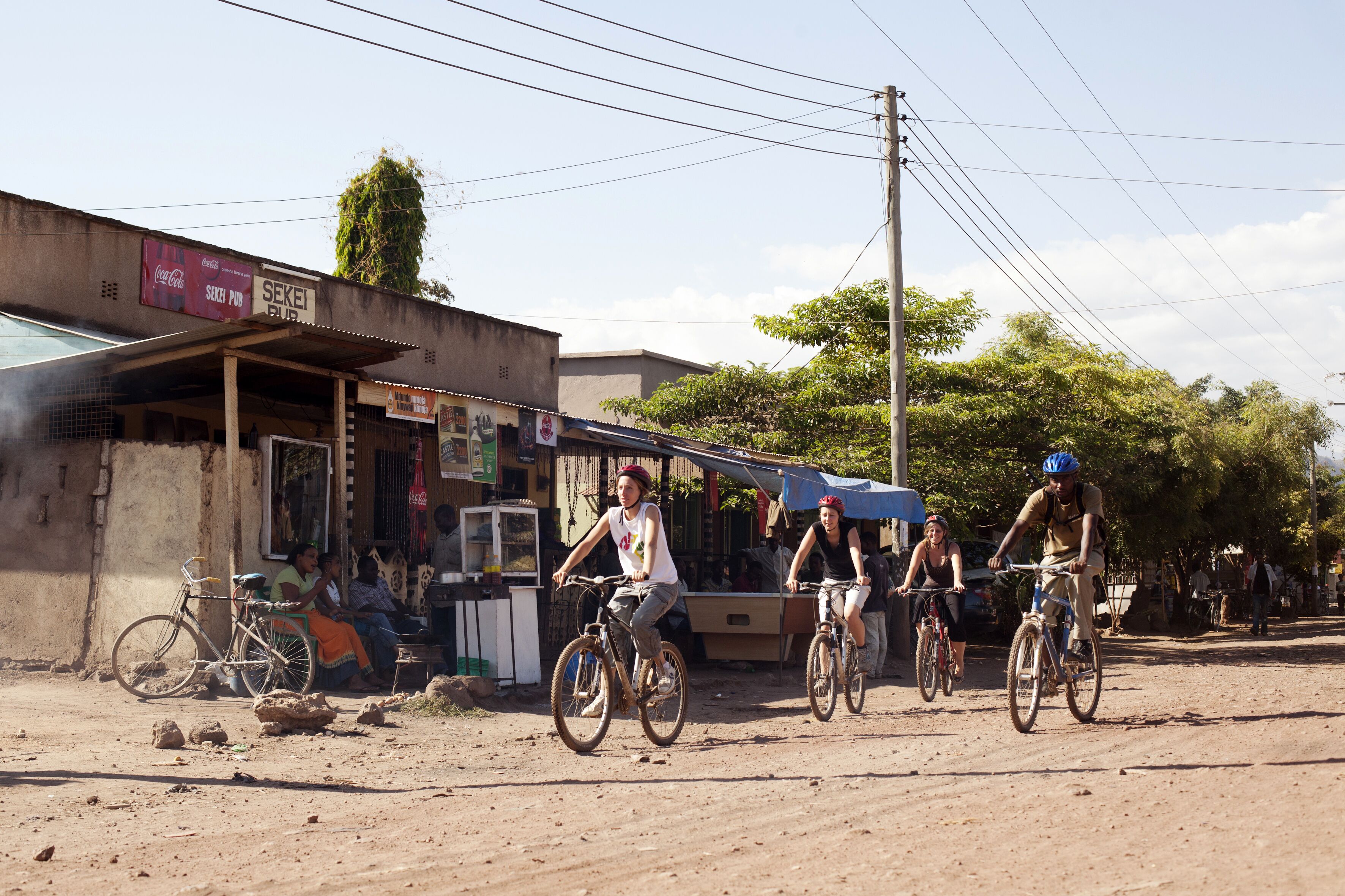 A group of people riding bicycles on a dirt road with buildings and a Coca-Cola sign in the background.