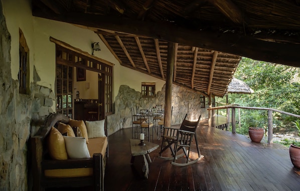 A rustic wooden deck with stone walls, a thatched roof, and a dining area with wooden furniture. - Rivertrees Country Inn (Arusha)