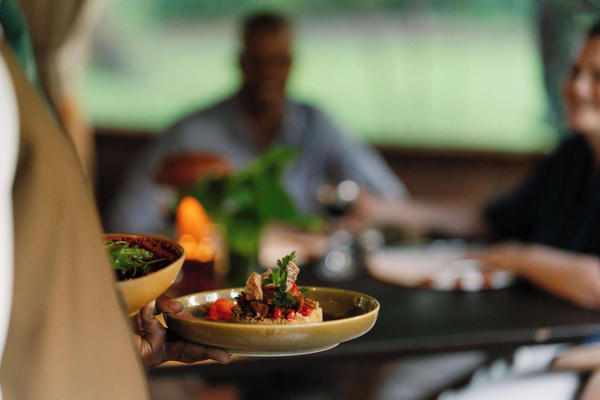 A person holding a plate with food, a table, and another person in the background.