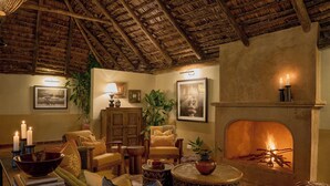 A cozy living room with a fireplace, wooden furniture, and a thatched ceiling. - Rivertrees Country Inn (Arusha)