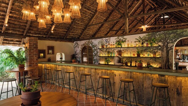 A bar area with wooden stools, a high ceiling with thatched beams, and a mural of tropical scenery. - Rivertrees Country Inn (Arusha)