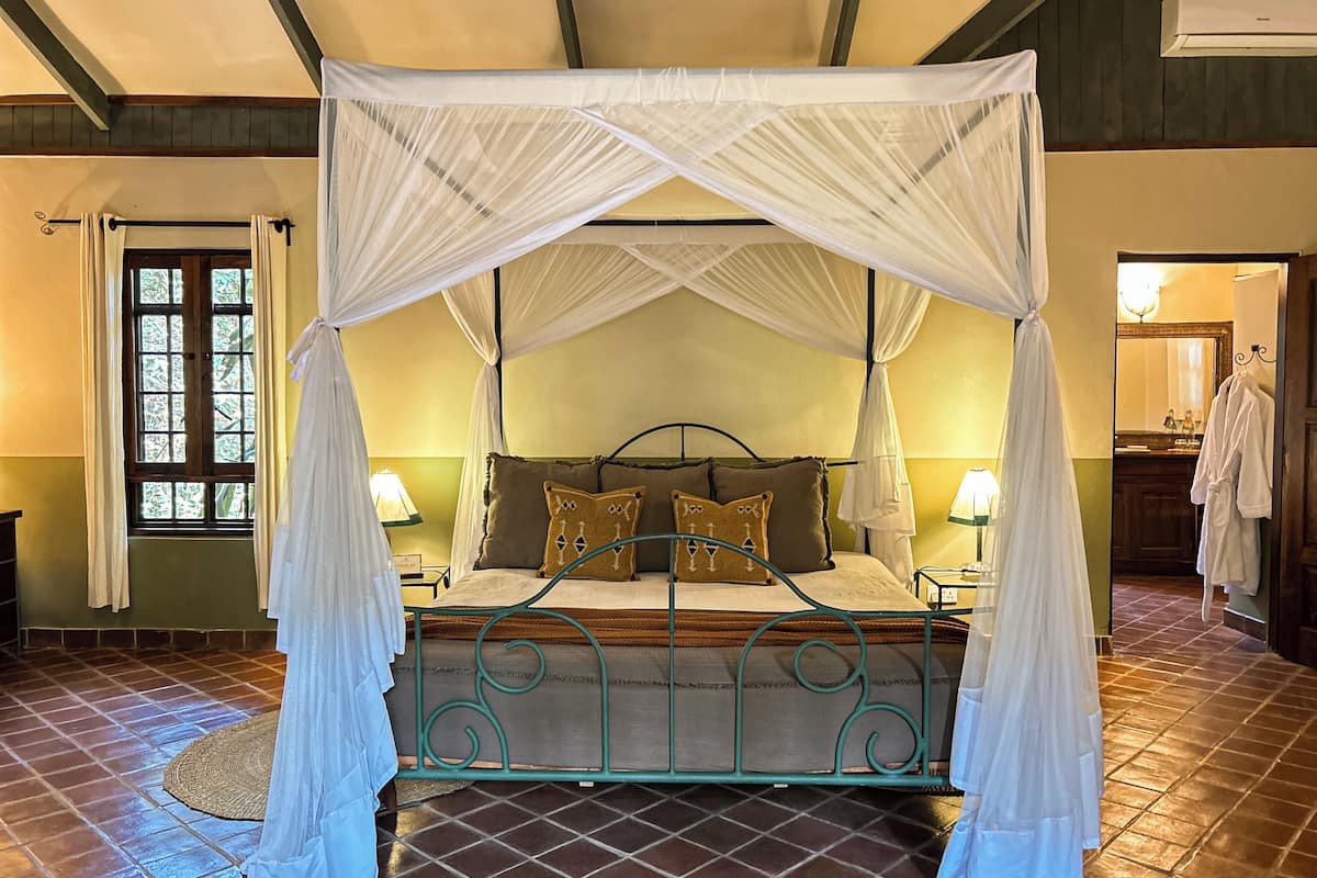A room with a bed under a canopy, a nightstand with a lamp, a window with curtains, and a bathroom with a mirror and towel rack.