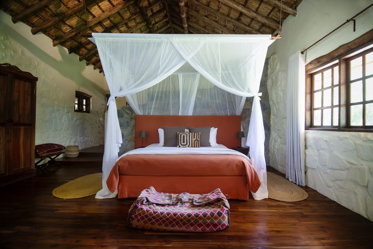 A bedroom with a four-poster bed, a stone wall, a wooden ceiling, and a window with curtains.