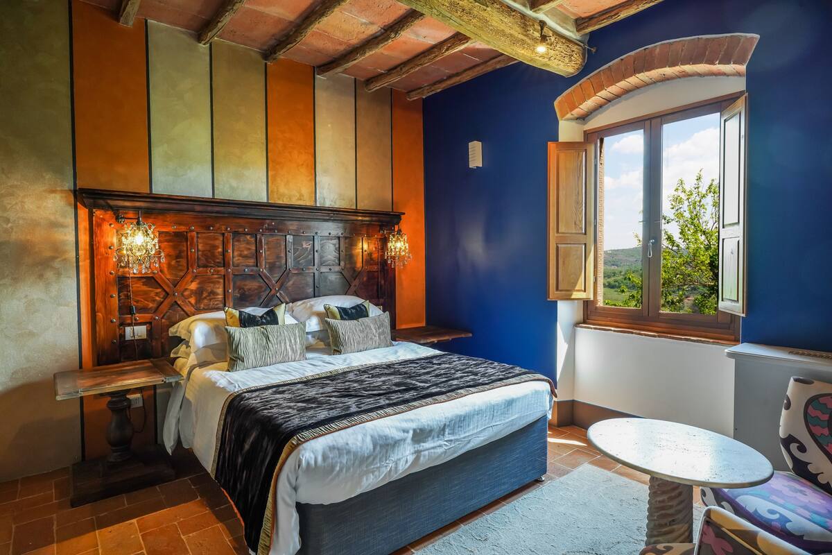 A bedroom with a large bed, a wooden headboard, a nightstand, a table, and a view of the outdoors through a window.