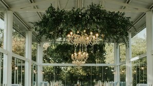 A long table set for a formal event, with clear chairs and white tablecloths, under a large chandelier and surrounded by greenery. - Il Verreno (Ambra)