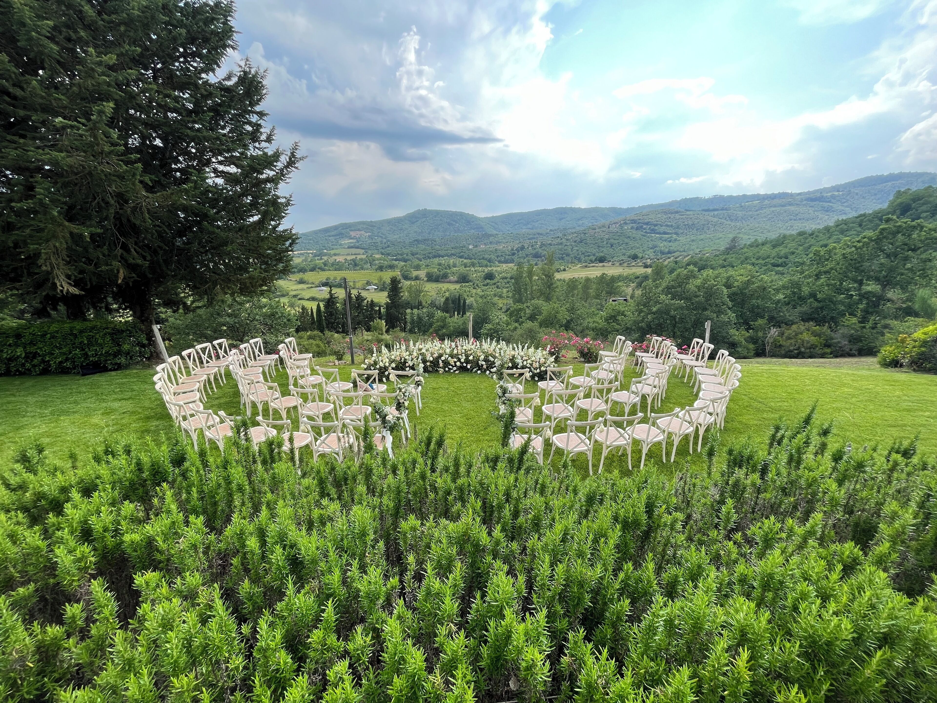A circular arrangement of white chairs in a grassy outdoor area with a backdrop of hills and trees.
