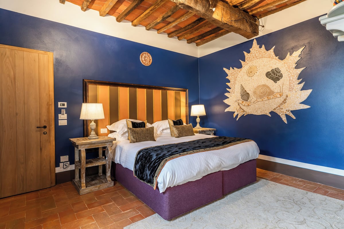 A bedroom with a large bed, wooden headboard, two bedside tables with lamps, a wall clock, and a decorative mosaic on the wall.
