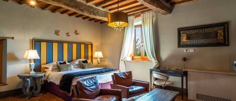 A bedroom with a bed, two leather chairs, a wooden table, a desk, and a hanging light fixture.