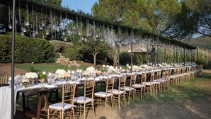 An outdoor dining area with long tables set for a meal, surrounded by greenery and trees. - Il Verreno (Ambra)