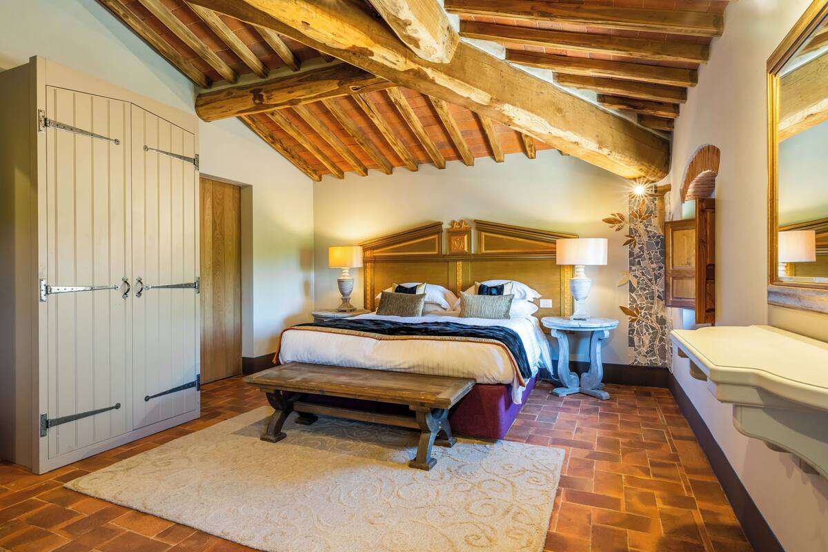 A bedroom with a wooden ceiling, a bed with multiple pillows, a bench, and a small table with lamps.