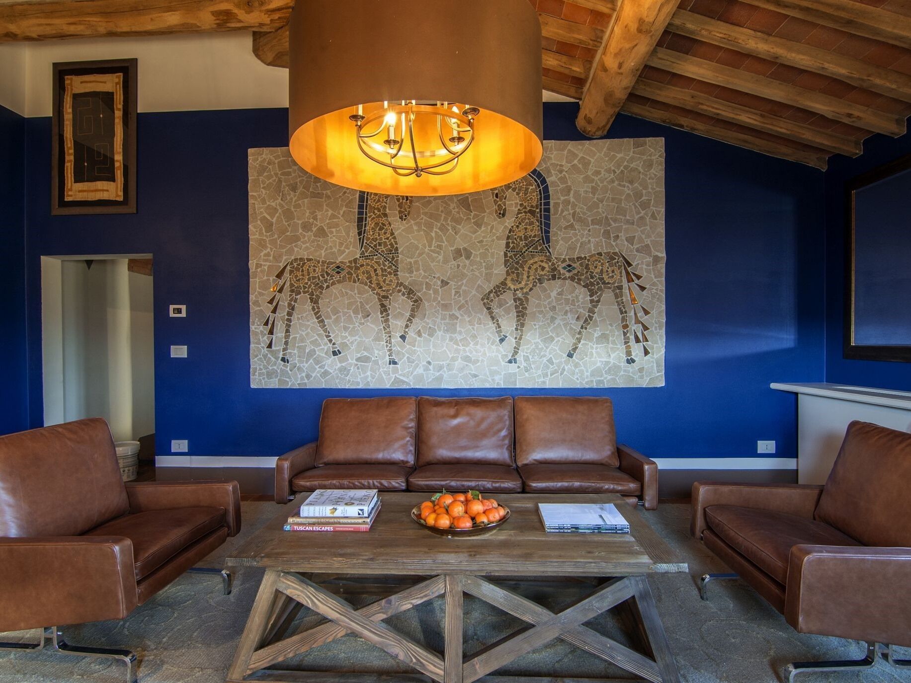A room with a brown leather sofa, a wooden coffee table with books and oranges, and a mosaic wall art of two horses.