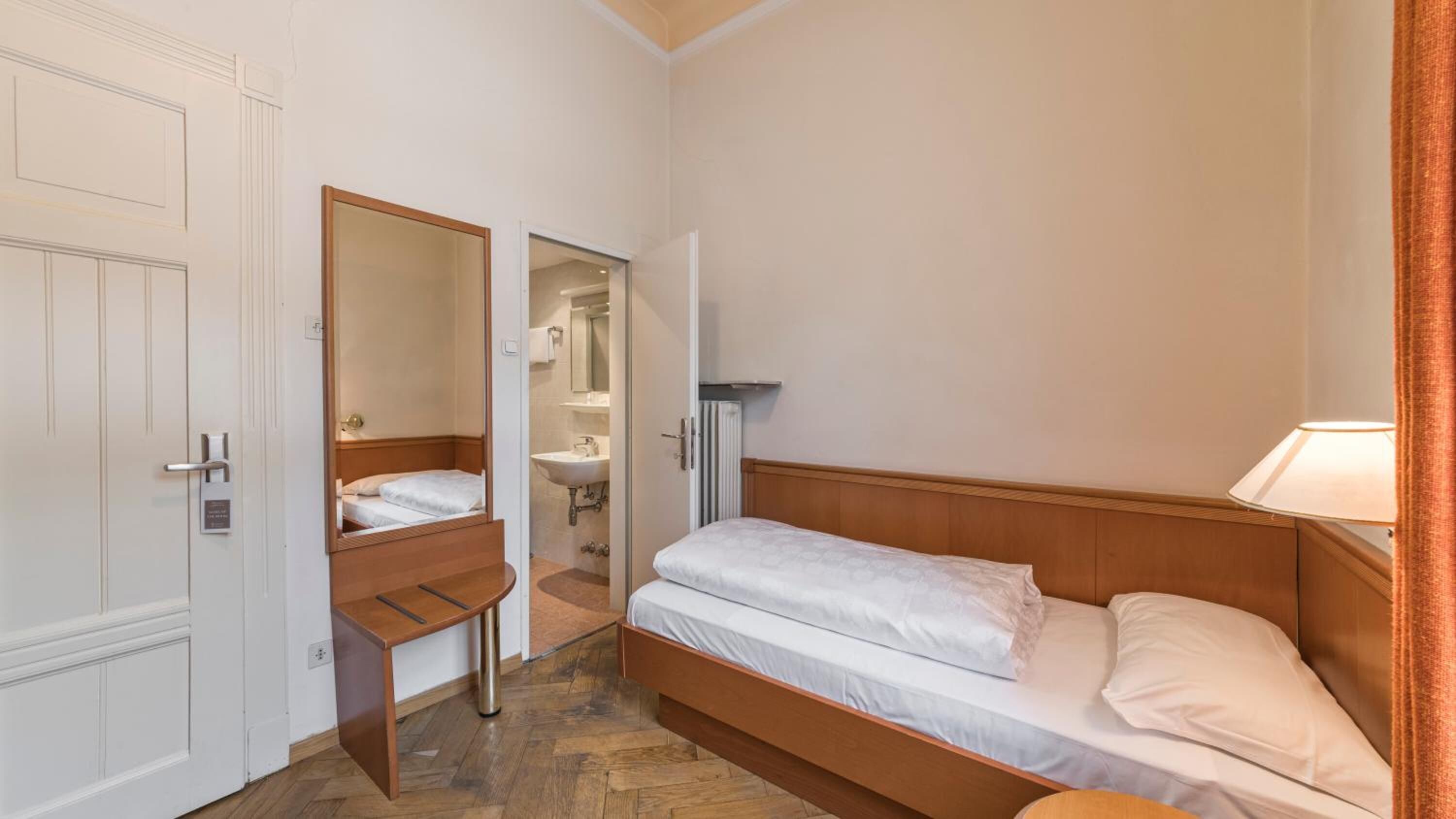 A small, compact bedroom with a single bed, a wooden nightstand, a mirror, and a door leading to a bathroom.
