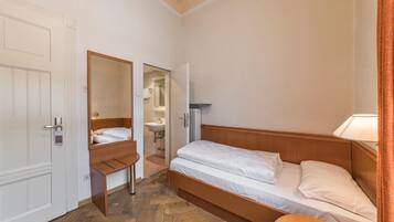 A small, compact bedroom with a single bed, a wooden nightstand, a mirror, and a door leading to a bathroom.